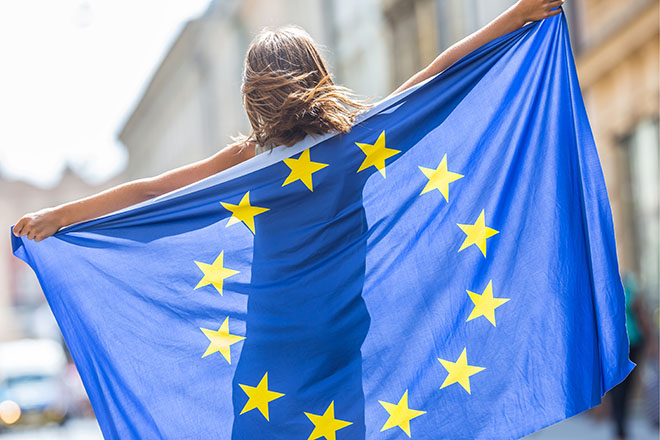 Mujer joven de espaldas sostiene una bandera de europea que la cubre casi por completo