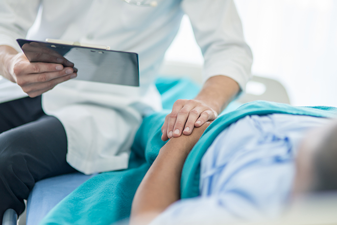 Paciente recostado y médico tocándole con su mano la mano del paciente mientras lo está visitando