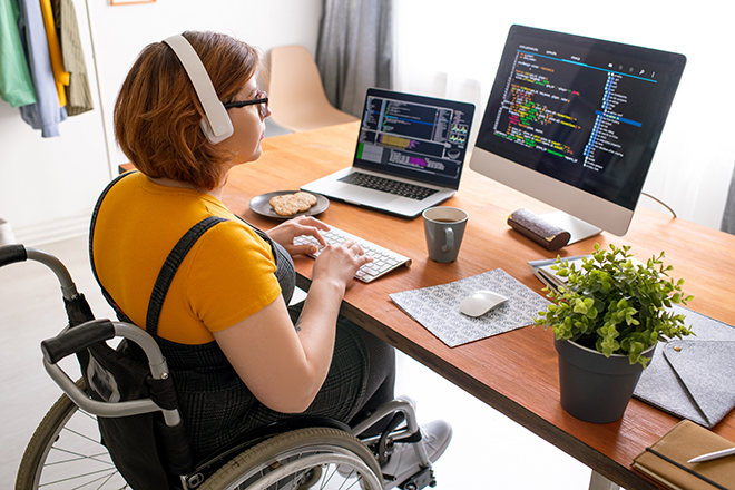 Programadora independiente en audífonos modernos sentada en silla de ruedas y usando computadoras mientras codifica juegos web en casa