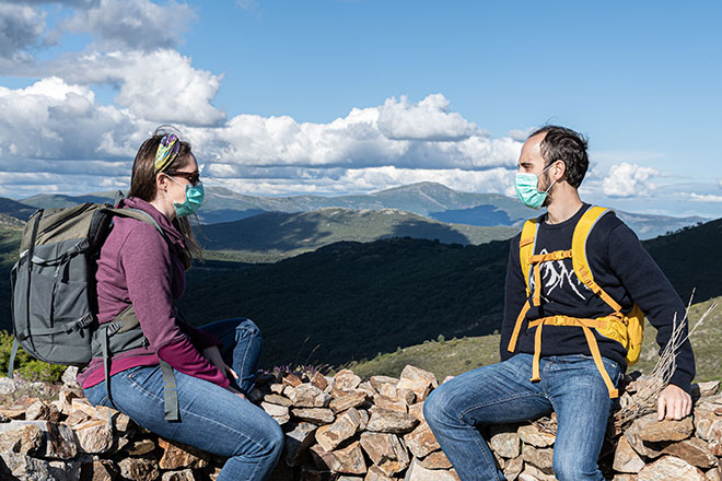 En un paisaje de montaña dos chicos con mochila y mascarilla quirúrgica están conversando.