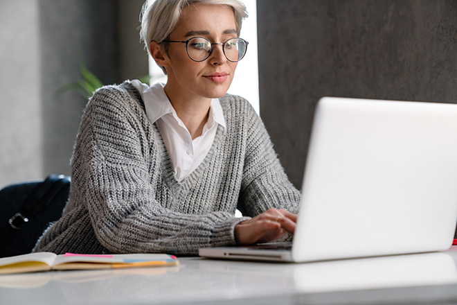 Mujer de pelo corto, cabello blanco y gafas en una mesa de oficina delante de un portátil blanco, y una agenda de papel