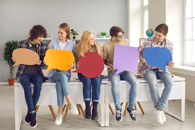 Cinco chicos jóvenes sentados en una mesa que sostienen con las manos una cartulina de colores sin nada en blanco.