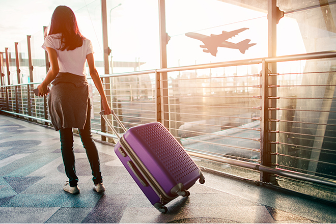 Mujer joven andando por un aeropuerto llevando una una maleta y a través de las vidrieras se ve un avión que despega
