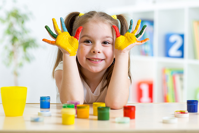 Niña en un aula, apoyada en un pupitre, con expresión alegre mostrando sus manos pintadas de colores