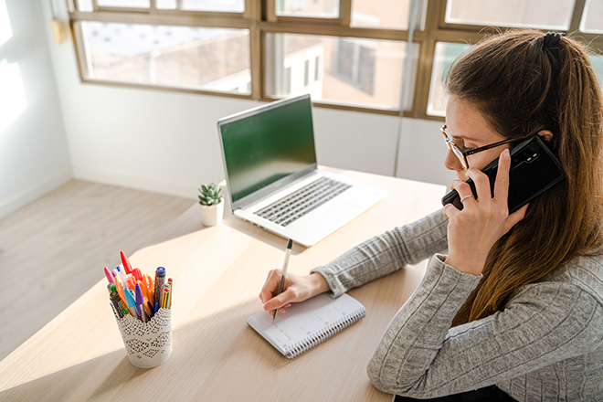 Mujer sentada en un escritorio, escribiendo en una agenda a la vez que hablar por teléfono móvil