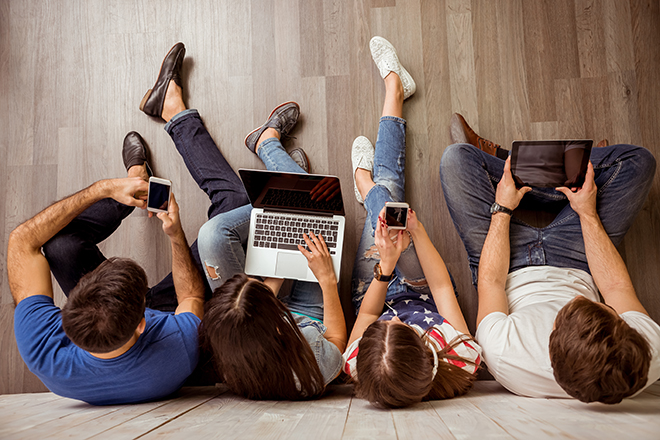 Vista cenital de dos chicas y dos chicos en el suelo apoyándose en la pared utilizando sus dispositivos electrónicos