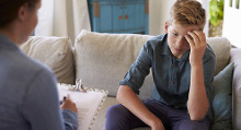 Una persona entrevista a un adolescente sentado en su sofá con la mano apoyada en la cabeza