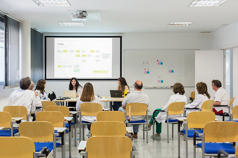 Una reunión en un aula. Una mujer joven está sentada en un escritorio frente a un ordenador. En la pizarra se proyecta la imagen de una página web y una aplicación móvil. 