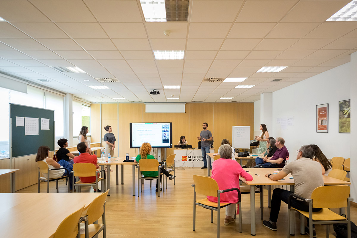 Una reunión en un aula, hay varios grupos de personas sentadas en mesa y tres mujeres y un hombre de pie están dirigiéndose al grupo